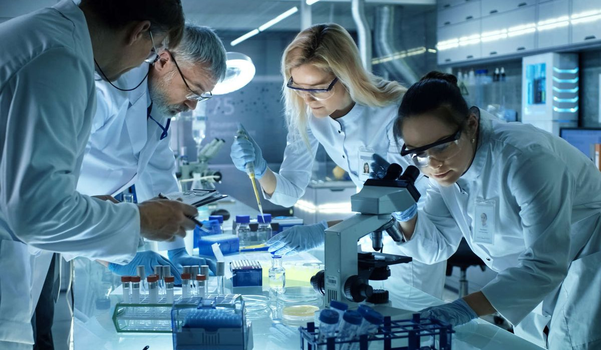 A group of scientists in lab coats and protective gear working together in a modern lab, performing research with test tubes, a microscope, and pipettes.