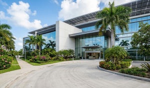 A wide-angle, hyper-realistic photo of the main entrance to a premium pharmaceutical facility in Puerto Rico, showing a modern glass and white concrete building with solar panels on the roof and tropical landscaping with palm trees.