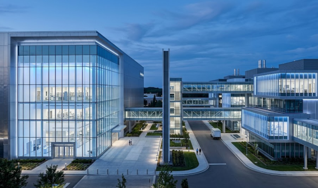 A high-angle photograph of a large, modern corporate or research campus at twilight. It features multiple interconnected glass and steel buildings. A prominent tower stands on the left. Elevated glass walkways connect the left buildings to a cluster on the right. Pedestrians walk on a paved plaza, and a white van is parked on an asphalt driveway. The sky is dark blue with clouds, and blue-tinted lights illuminate the interiors and paths.