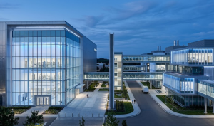 A high-angle photograph of a large, modern corporate or research campus at twilight. It features multiple interconnected glass and steel buildings. A prominent tower stands on the left. Elevated glass walkways connect the left buildings to a cluster on the right. Pedestrians walk on a paved plaza, and a white van is parked on an asphalt driveway. The sky is dark blue with clouds, and blue-tinted lights illuminate the interiors and paths.